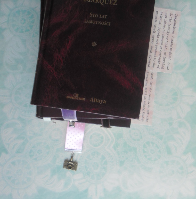 Trzy książki w ciemnych okładkach ułożone w stos, z których wystają zakładki wstążkowe: różowa w kropki z aparatem fotograficznym, miętowa i brązowa z walizkami i fioletowa z wieżą Eiffla.